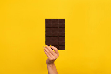 Female hand with yellow nail polish holding a dark chocolate bar on vibrant yellow background. Minimalist composition with copy space.
