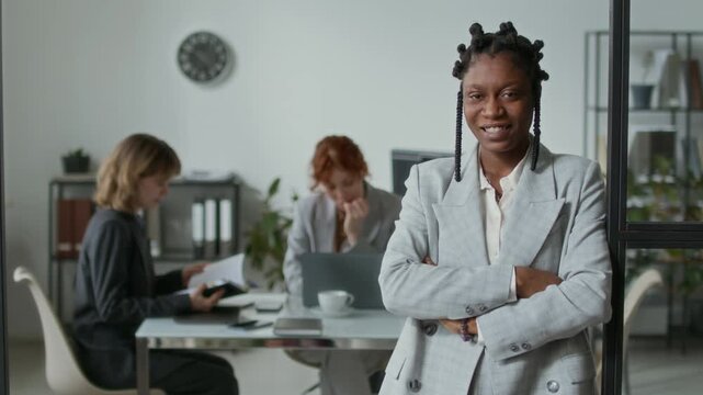 Waist-up portrait of African American young businesswoman dressed in white shirt and grey jacket smiling for camera with folded arms while two female coworkers sitting at office table in background
