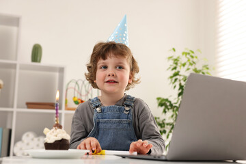 Birthday celebration. Little boy with cupcake having video call via laptop at table indoors