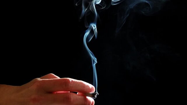 Close-up of a hand holding a cigarette with smoke rising against a dark background