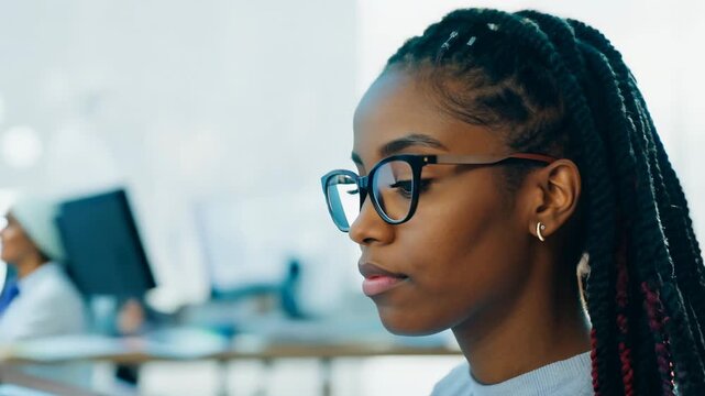 Young African American woman with multi colored dread locks in front of laptop monitor in office. A moment of determination shines as she tackles challenges in her workspace.
