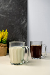 Choice of morning beverage: a glass mug of fresh milk or black coffee. Yellow Solidago flowers in the background against a modern textured wall.