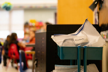 Close-up detail of educational materials and student supplies neatly organized on rolling cart inside elementary school classroom with young students sitting in small chairs visible in soft focus back © Jeanne Sager Photo