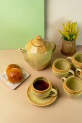 Afternoon tea time with a ceramic teapot, a cup of black tea, empty cups, and bread rolls. A cozy and elegant tableware setup with flowers.