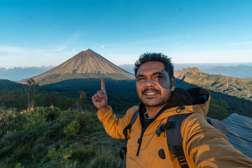An Asian traveler in a yellow jacket points at Mount Inerie volcano in East Nusa Tenggara, Indonesia. A scenic adventure shot under clear blue sky, ideal for travel and nature themes.   © Ryan