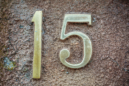 Polished brass house number 15 mounted on a rough, cracked reddish-brown wall. The elegant, slightly curved number style contrasts with the textured surface, giving a vintage yet refined appearance