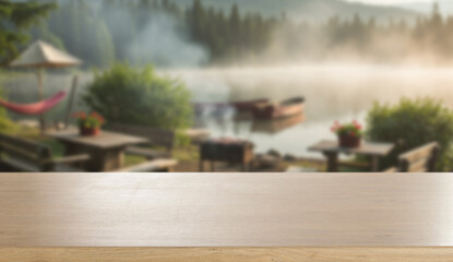 Wooden table with space for product in front of barbecue by the lake – summer camping and outdoor cooking concept. Blurred background of calm lake with distant mountains and a clear blue sky.