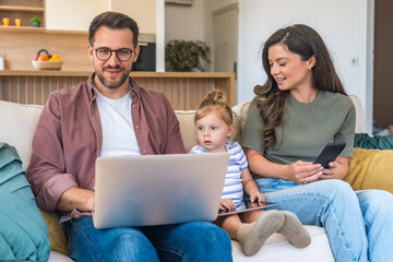 Fototapeta premium Young happy business family. Businessman and businesswoman trying to work at home online on laptop computer, but their little baby girl dont let them, wanted to play and have fun. Freelance parents.