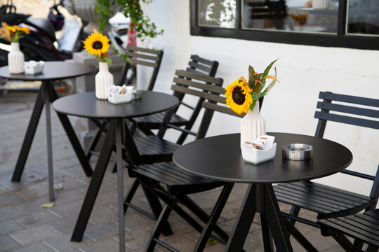 Outdoor cafe terrace with minimalist black tables, sunflowers in white vases, and empty chairs. Cozy modern design on a city sidewalk, perfect for relaxation or coffee break visuals.