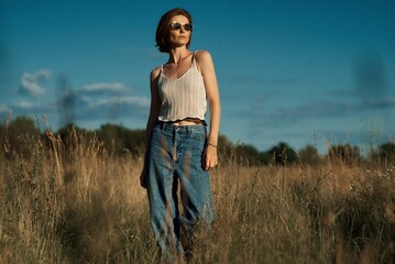 A woman in a field poses wearing sunglasses with a beautiful blue sky.