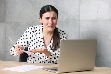 Young businesswoman with papers and laptop complaining to someone via phone at wooden table indoors