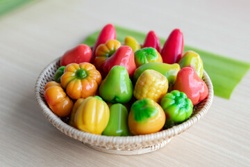 Colorful Thai mung bean desserts (look choop) shaped like mini fruits and vegetables, displayed beautifully in a wicker basket on a wooden table. Traditional Thai sweet.