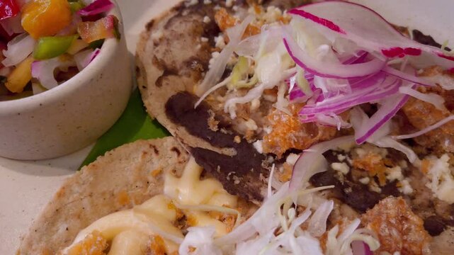 Close-up of a savory Oaxacan tlayuda layered with black beans, cheese, fried pork skin, radish, pickled onions and lettuce, served with a side of spicy salsa