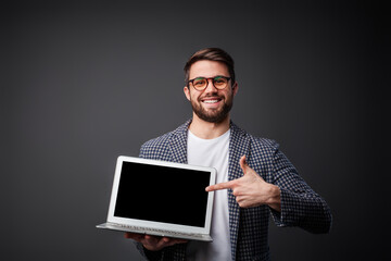 A cheerful man is presenting a laptop with a blank screen, wearing glasses and a stylish outfit, standing against a plain dark background, emanating professionalism and a positive, approachable vibe.