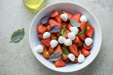 Plate of strawberry caprese with cherry tomatoes, mini mozzarella and basil, horizontal shot on a beige stone background, elevated view