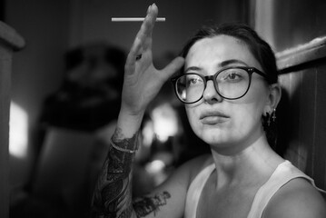 A pensive woman with glasses holds a cigarette in a black and white photo.
