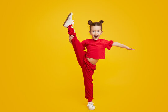 Adorable little girl in bright outfit lifting leg and smiling while dancing against yellow background. Dancing class for kids concept