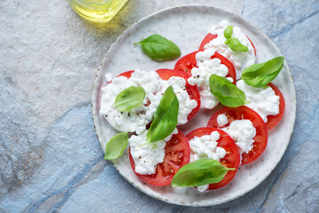 Plate of cottage cheese caprese with ripe tomatoes and green basil, horizontal shot on a blue and beige stone background, elevated view