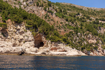 Natural cave in rocky cliff surrounded by pine trees and shrubs, seen from the sea. Rugged...