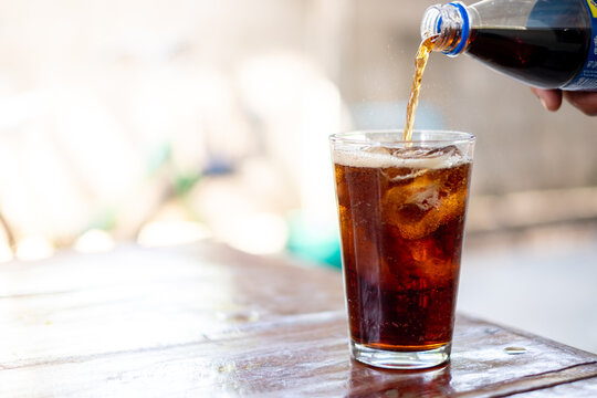 Refreshing cola soda being poured into a glass with ice cubes on a wooden table, perfect for beverage advertising, summer themes, and food and drink promotions.
