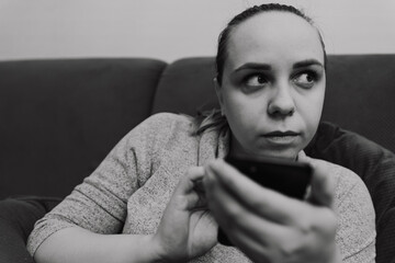 A woman sits on a couch holding a smartphone with a worried expression She appears to be thinking...