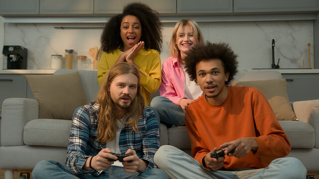 Four multiracial people friends students fun weekend at home Caucasian men guys males playing console video game addicted win lost African American women girls females support laughing joy friendship