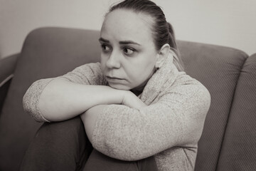 A woman sits on a couch hugging her knees and gazing off into the distance with a thoughtful...