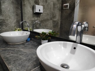 Fototapeta premium A modern bathroom counter features two white vessel sinks and chrome faucets against a dark marble wall with a large reflective mirror.