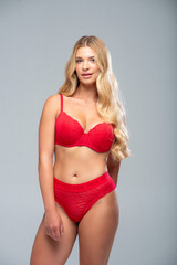 Fashion-style portrait of a slim blonde female model in red underwear, posing against a soft grey...