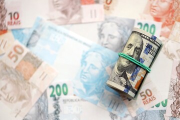 Roll of US dollar bills lies on big amount of Brazilian reais banknotes close up