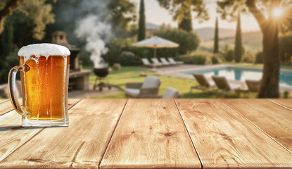 Glass of beer on wooden garden table – backyard relaxation at home. In the blurred background, a lush green garden and patio area with outdoor furniture behind a house.