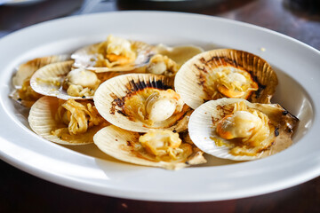 Grilled scallops in the shell with butter, a fresh seafood menu of a restaurant, served on a white plate on the wooden table with Thai seafood sauce.