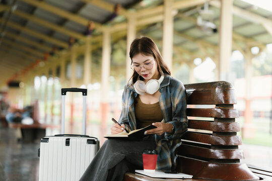 A young woman with glasses and headphones sits on a bench at a train station, holding a smartphone and looking around, surrounded by luggage and coffee.