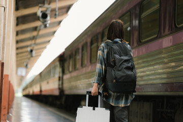 A young woman walks through a train station with a suitcase, headphones around her neck and looking at her phone, ready for a trip or a journey.