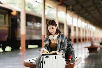 A young woman with glasses and headphones sits on a bench at a train station, holding a smartphone...