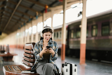 A young woman with glasses and headphones sits on a bench at a train station, holding a smartphone...