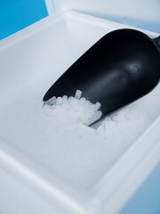Black plastic scoop filled with white dry ice pellets inside white styrofoam container on blue...
