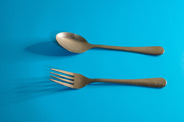 A spoon and a fork are placed on a blue surface, casting shadows.