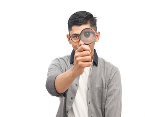 Young man holding magnifying glass in front of eye with curious expression and wearing glasses on white background