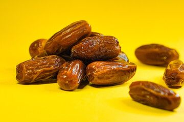 A small pile of dark brown dates rests on a vibrant yellow background, a few others scattered nearby.