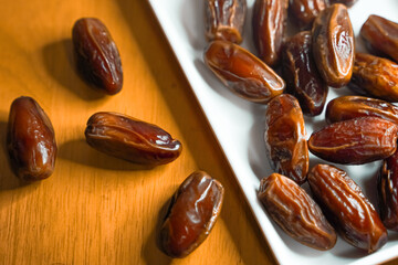 A close-up view of several dark brown dates, some scattered on a wooden surface and others neatly arranged on a white rectangular plate.