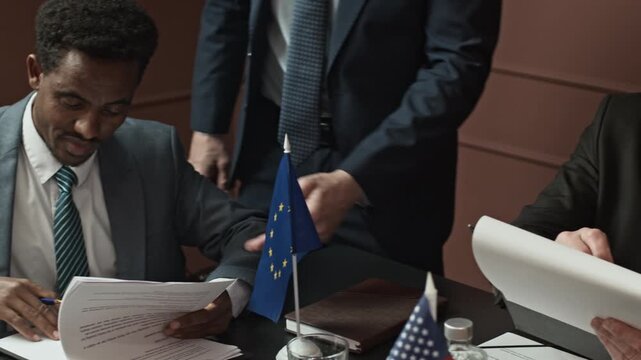 Pan shot of attache in formal suits guiding diverse delegates signing papers during international meeting at table