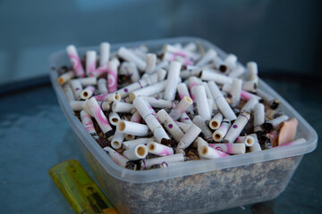 Close-up of a plastic container filled with numerous used cigarette butts and ashes, highlighting tobacco addiction, pollution, and environmental waste issues.