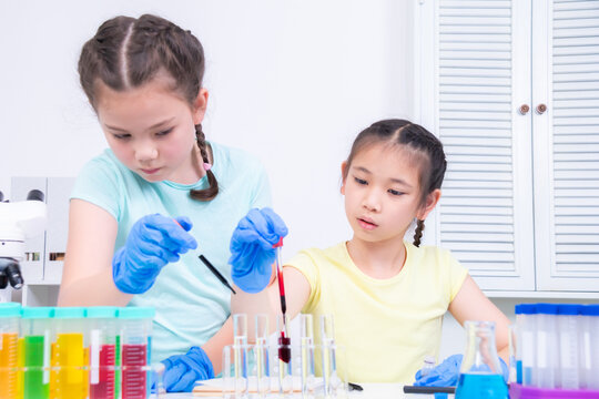 Two students wear gloves and drop colored liquid into a test tube, focus, collaborate to observe reactions in laboratory. Academic schoolgirls explore chemistry using glassware, curiosity children