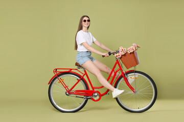Smiling woman in sunglasses riding bicycle with basket of flowers on green background