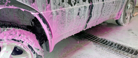 Car wash close-up with white and vibrant pink foam covering the vehicle. Vehicle Cleaning process with pink detergent. Self-service station, no people. For auto care, maintenance, service concepts