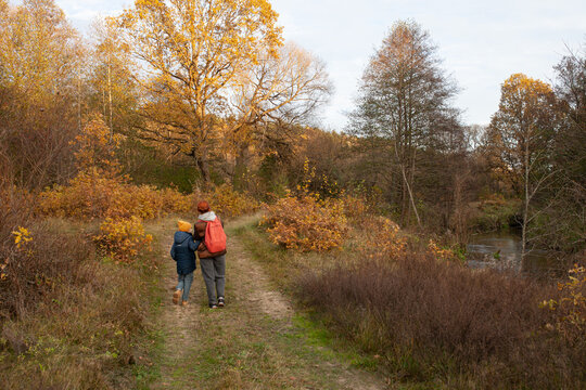 Back view of little boy and woman with backpack walking along river in autumn. Concept of ecotourism, hiking, travel with children, connection with nature.