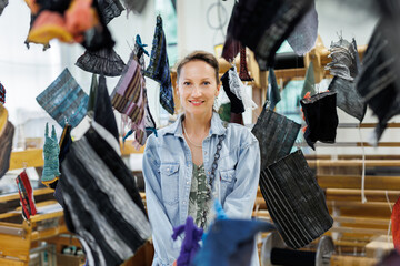 Woman standing in textile workshop surrounded by suspended fabric samples. She smiles gently while...