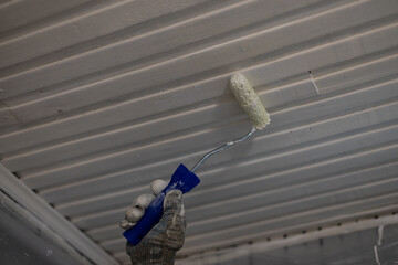 Gloved hand using a roller brush to apply white paint on a wooden ceiling, showcasing a home...