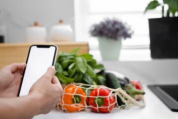 Hand with white screen phone near fresh vegetables and fruits in a reusable bag in the modern kitchen. Food delivery. Grocery shopping online. Eco-Friendly Shopping. The idea of a healthy lifestyle